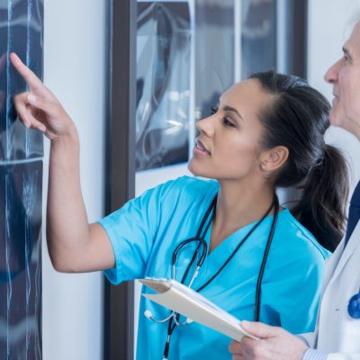 Mid adult female radiologist points to something on an x-ray image. A senior male doctor is also reviewing the x-ray. He is holding the patient's medical chart.