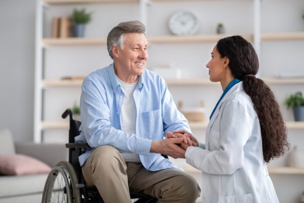 Older people healthcare support concept. Doctor holding hands of disabled senior man in wheelchair during medical visit. Portrait of handicapped eldelry male and medical specialist at home
